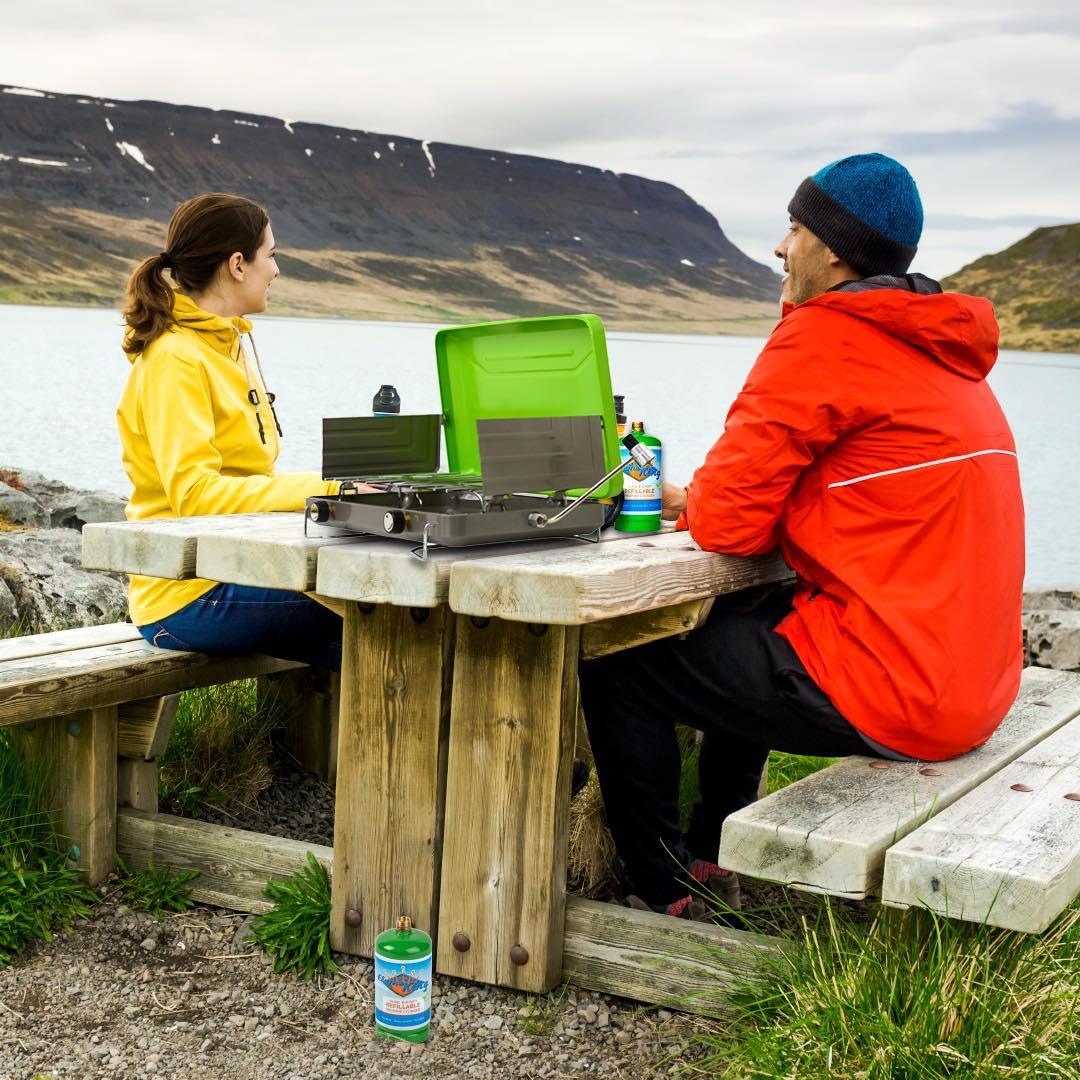 Two people at a lakeside picnic table using a portable propane camping stove outdoors