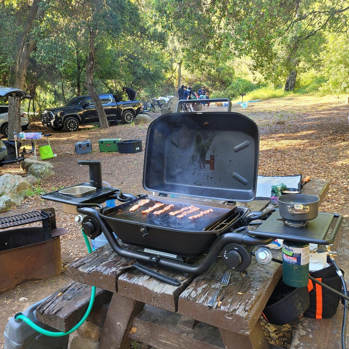 Portable outdoor grill cooking bacon at a wooded campsite with overlanding gear nearby