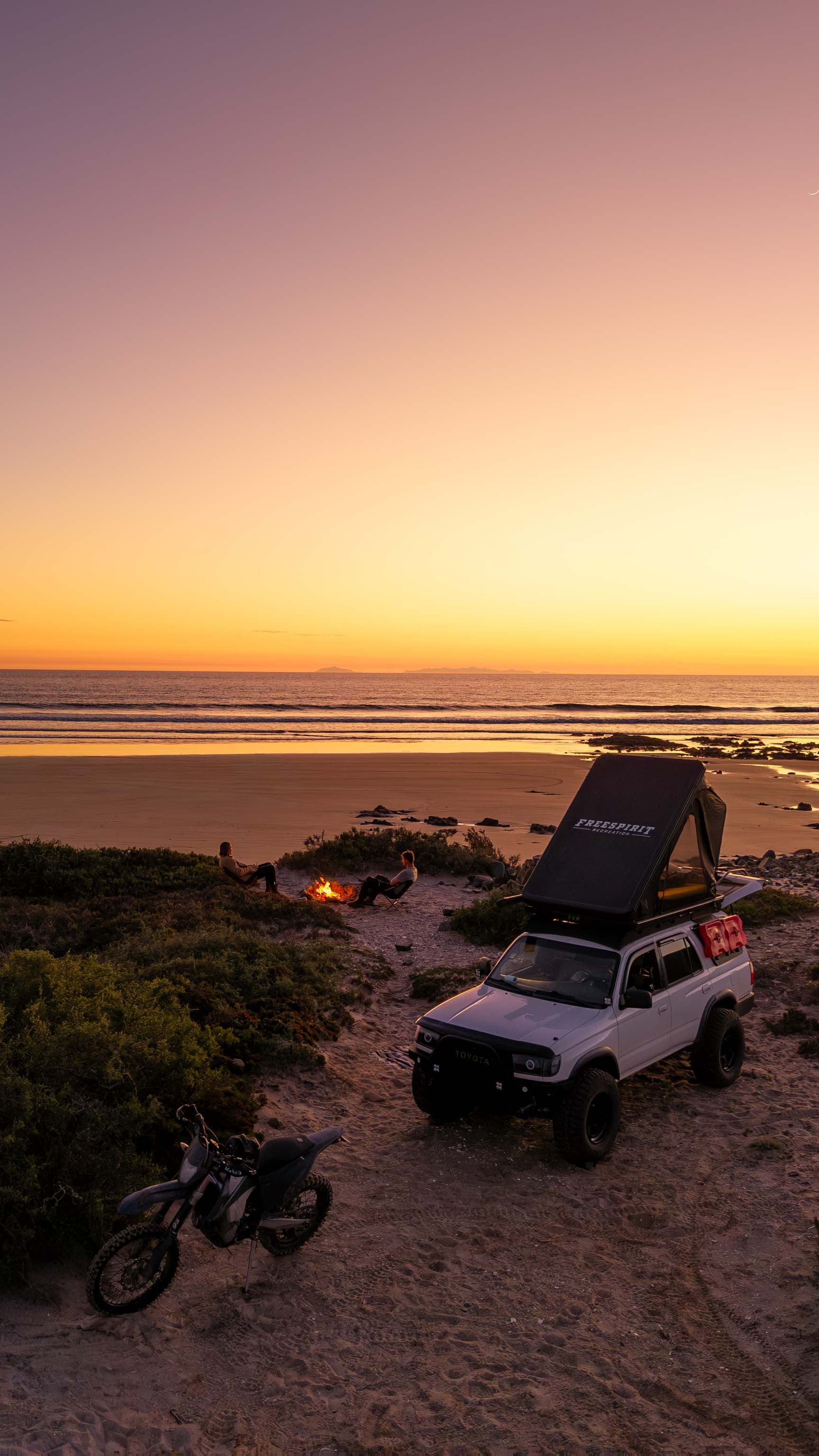 Overland SUV with rooftop tent and dirt bike on sandy beach at sunset, campfire nearby