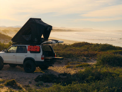 SUV with rooftop tent and open storage drawer parked on a beach at sunset