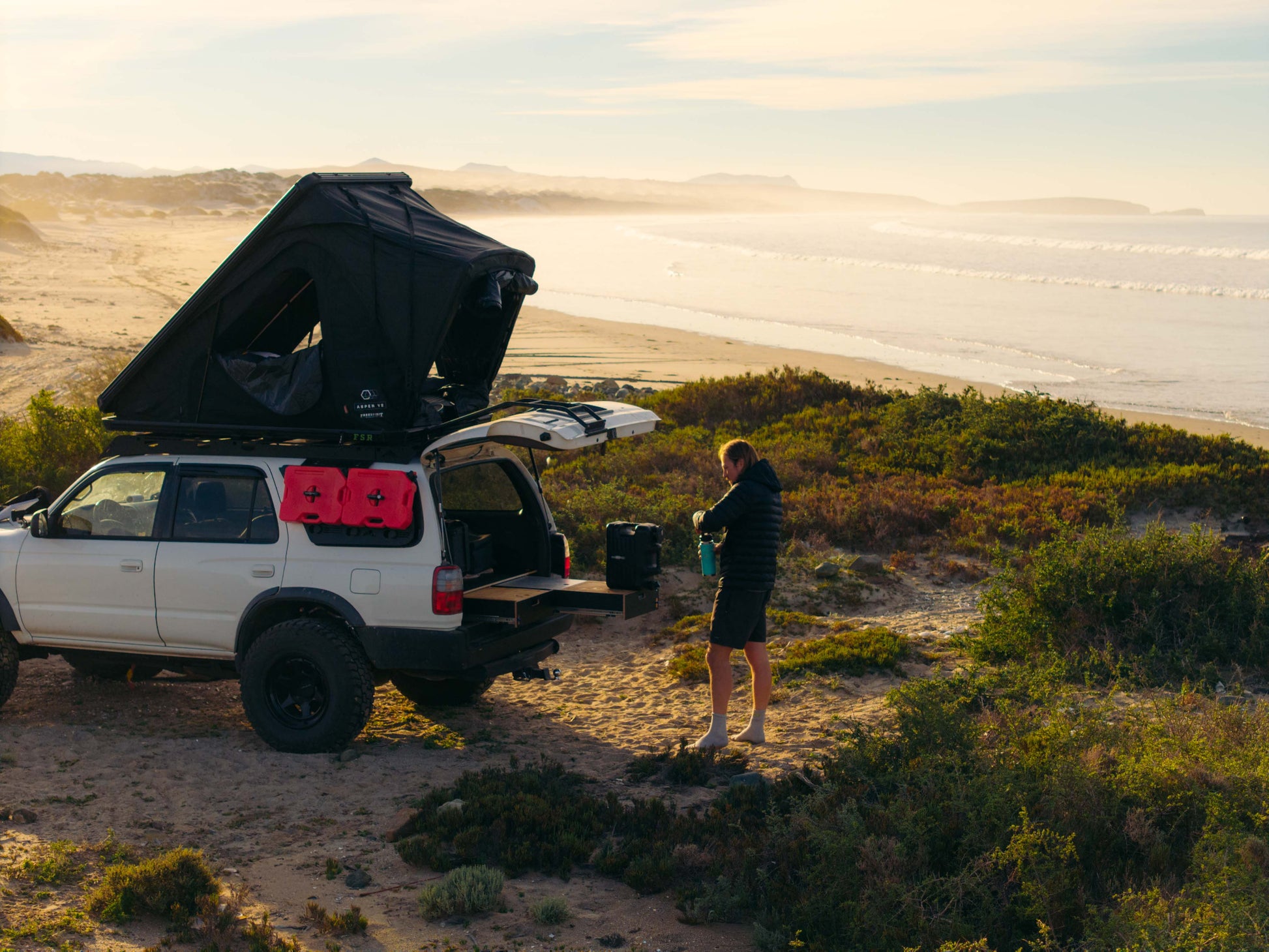 SUV with rooftop tent, vehicle drawer system, and camping gear on a beach at sunrise
