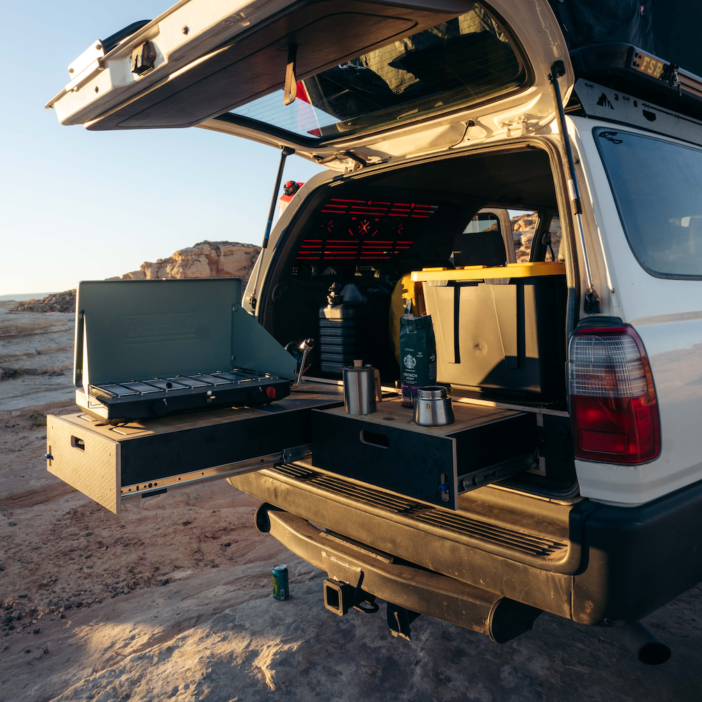 A white SUV parked in a desert canyon with its rear hatch open, showing a mounted outdoor kitchen setup with a portable stove, storage drawers, and camping gear inside and beside the vehicle.
