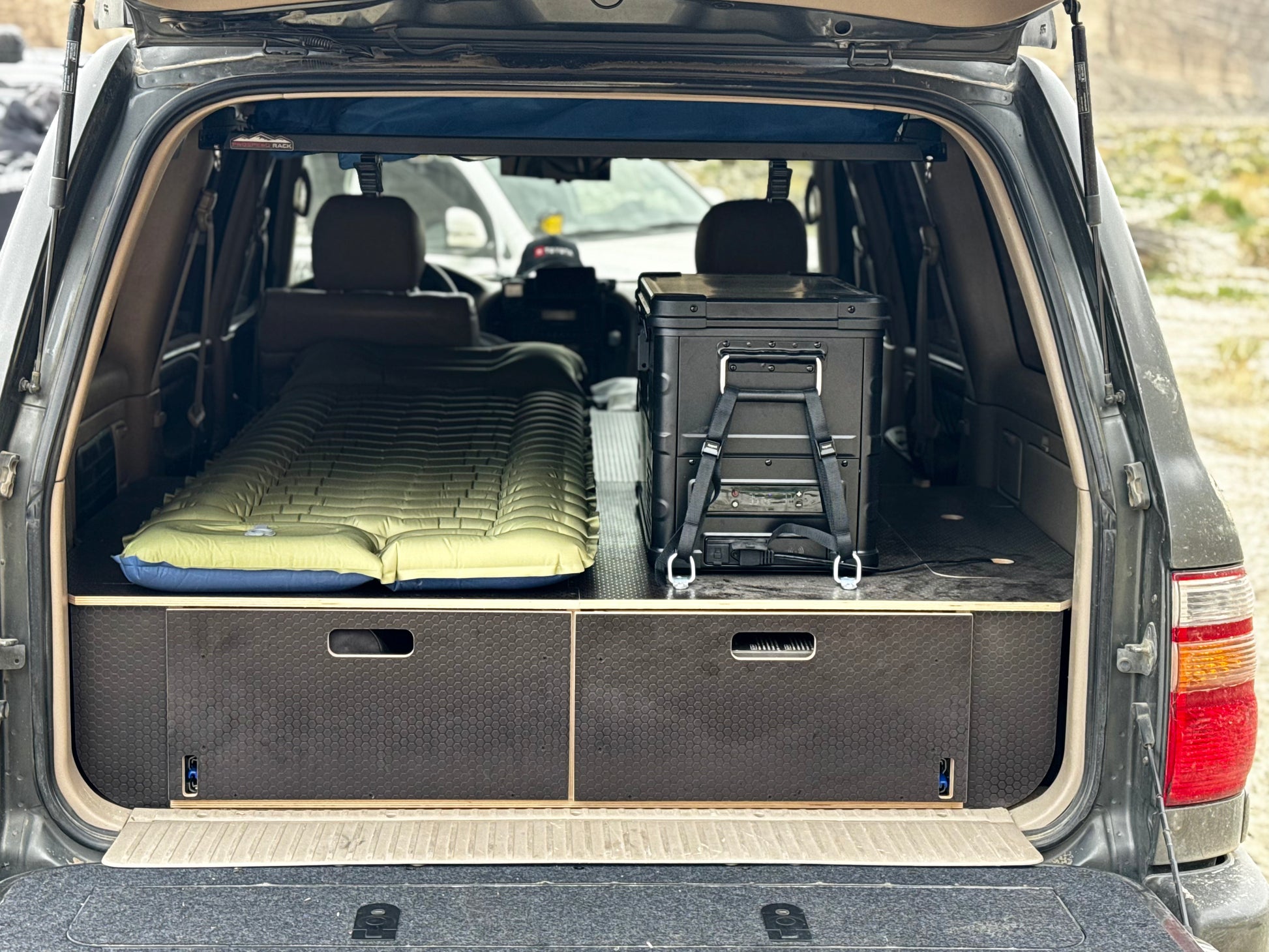 SUV trunk with custom wooden drawer system, green camping mattress, and storage box.