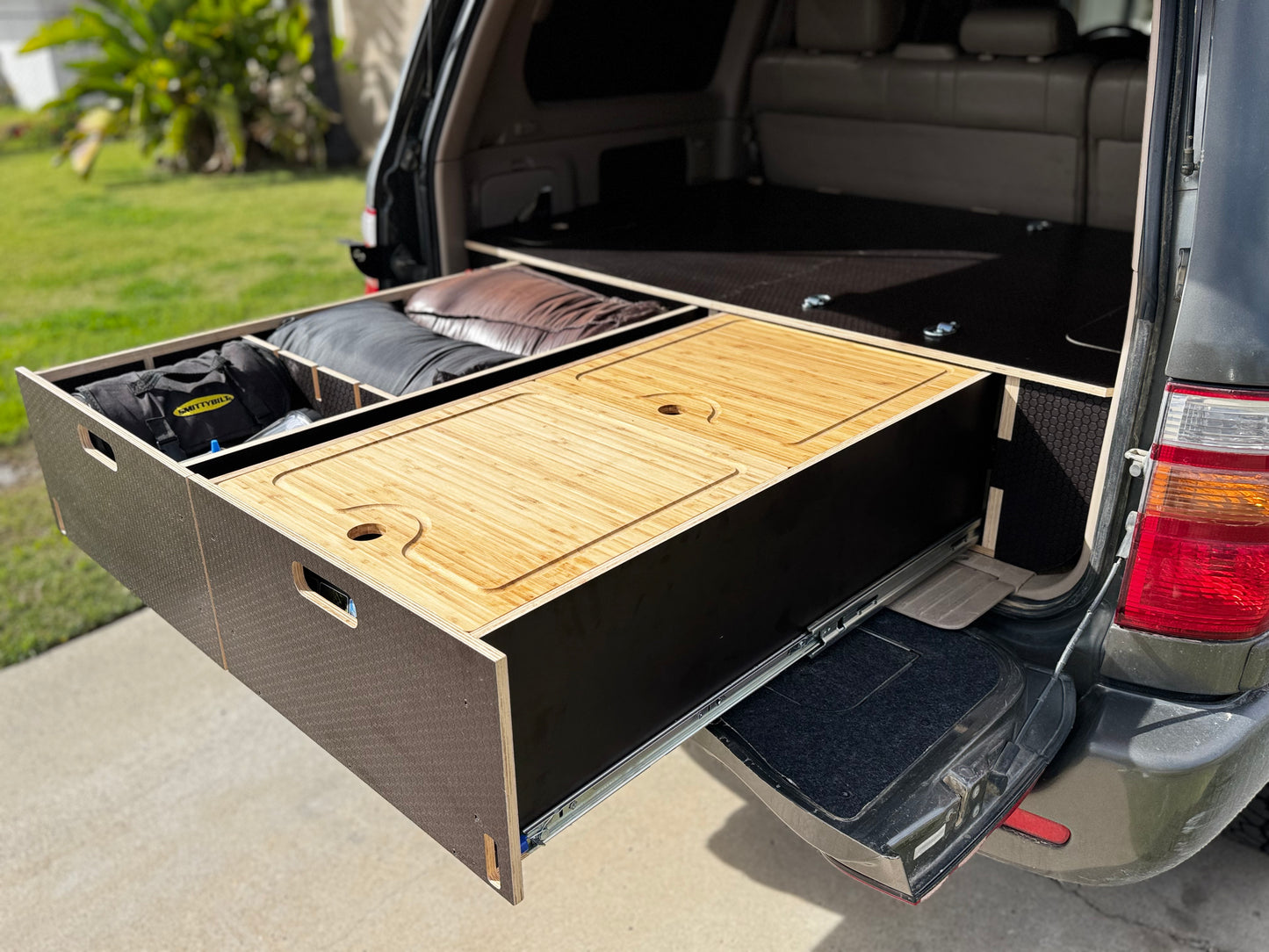 Solid wood vehicle storage drawer system installed in SUV trunk, organized with gear and bedding.