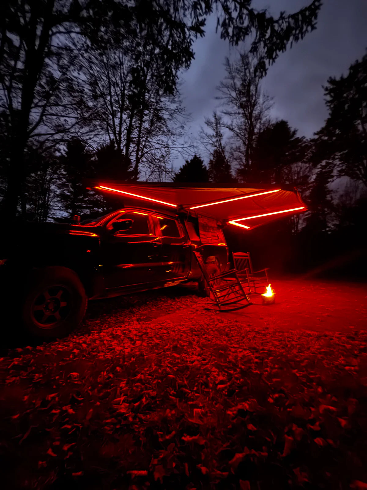 Truck with LED awning lights at campsite, two chairs and campfire in the woods at night