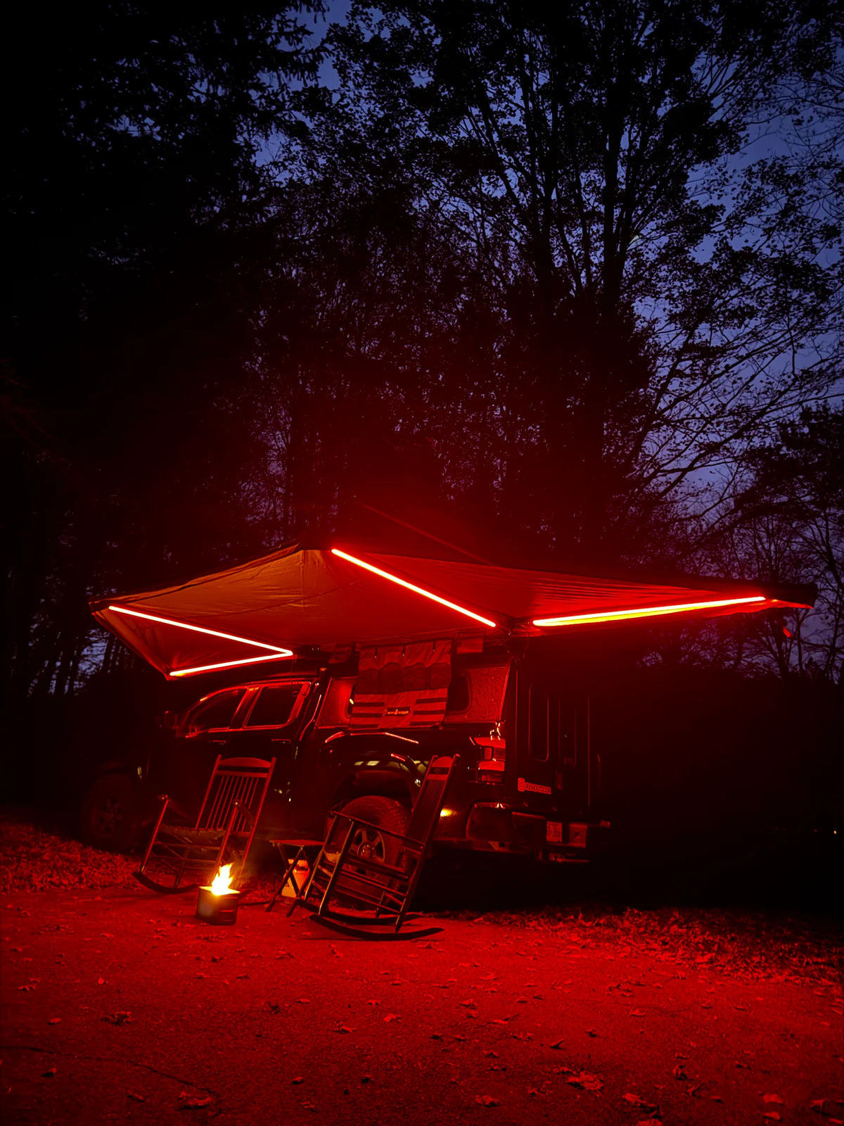 Truck with awning and red LED lights parked in forest campsite at night, rocking chairs by campfire.