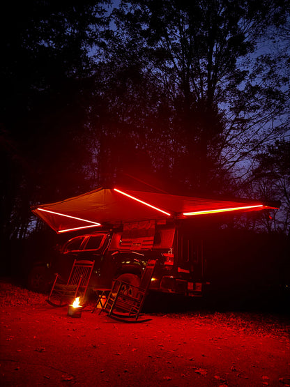 Truck with awning and red LED lights parked in forest campsite at night, rocking chairs by campfire.