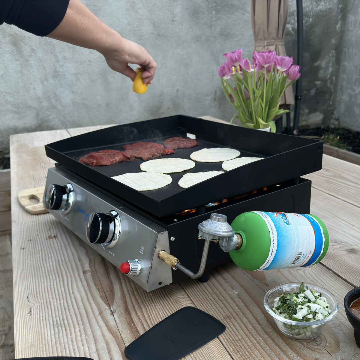 Portable propane camping griddle cooking meat and tortillas on a wooden table