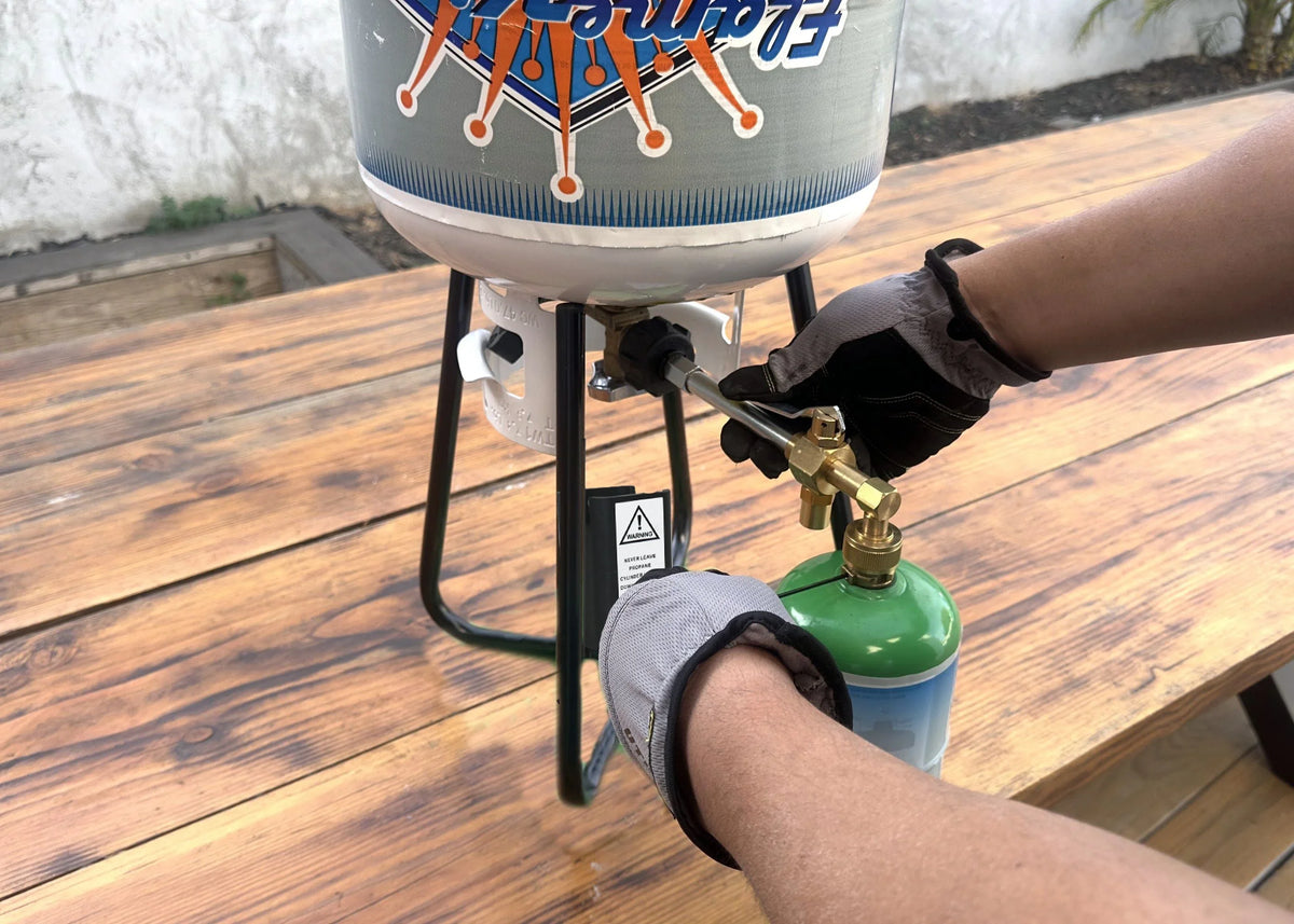 Hands in gloves connecting a small green gas canister to a portable outdoor cooker.