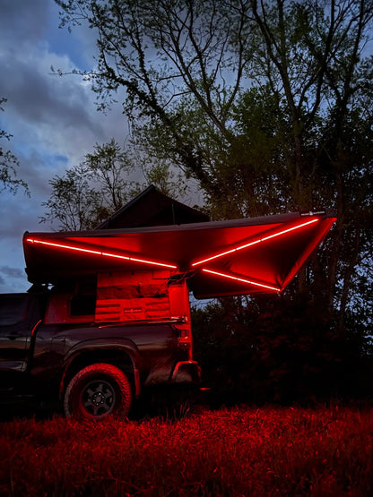 Truck with red LED-lit rooftop tent and awning in outdoor camping setting at dusk